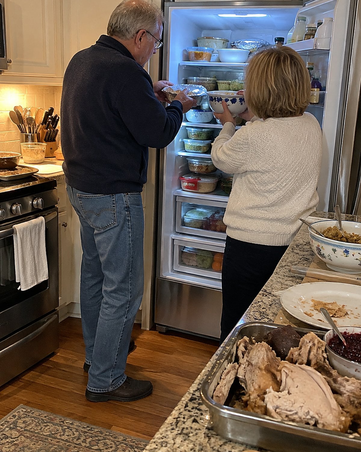 A couple in their sixties at the open fridge door the day after Thanksgiving, evening stove light on their hands as they shift foil-covered dishes across full shelves, nobody posing.