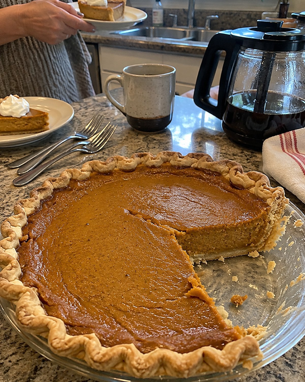 A partly cut pumpkin pie waiting beside a coffee pot and scattered dessert forks.