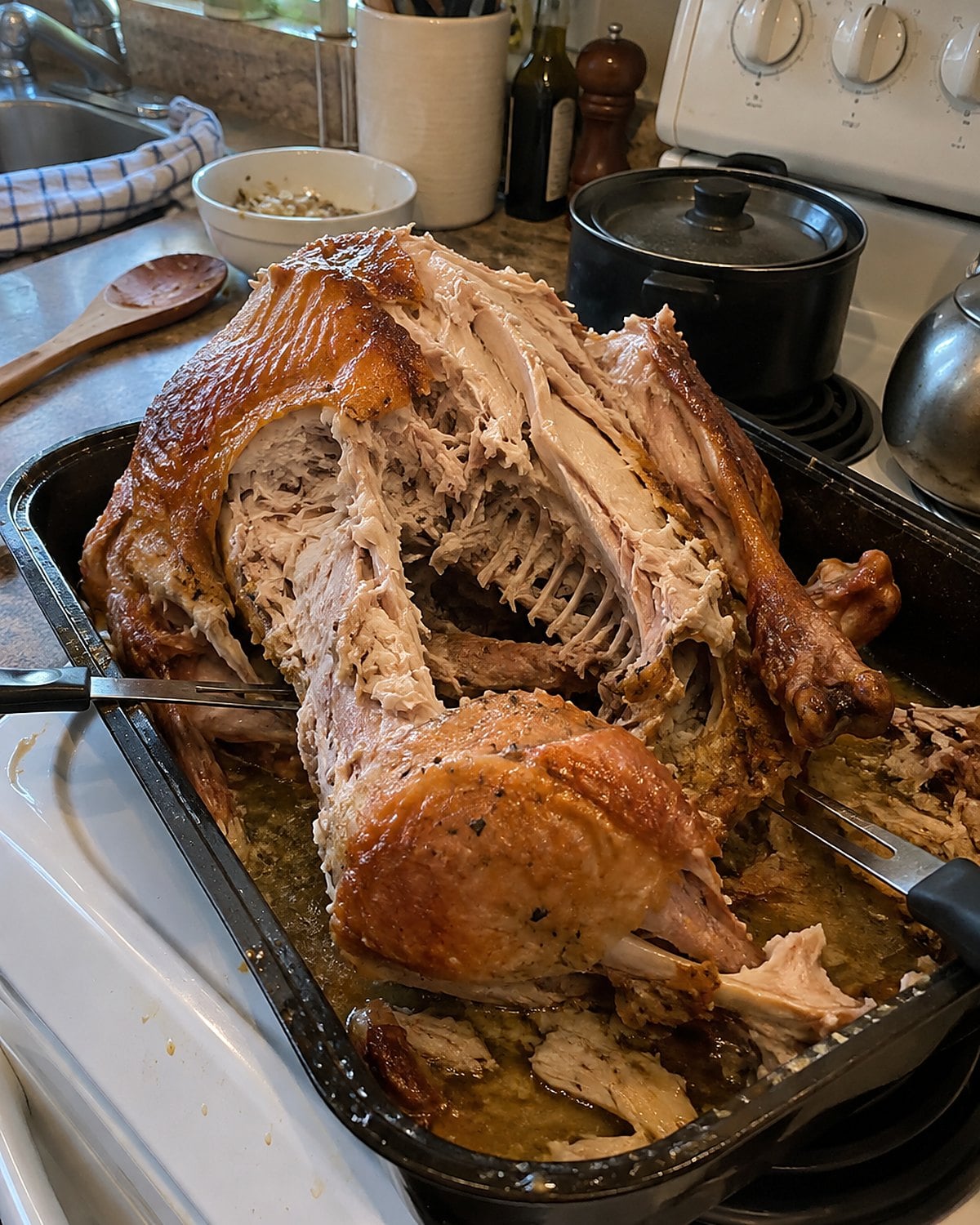 An older womanโs hands lifting a turkey carcass from a roasting pan beside a crowded white stove.