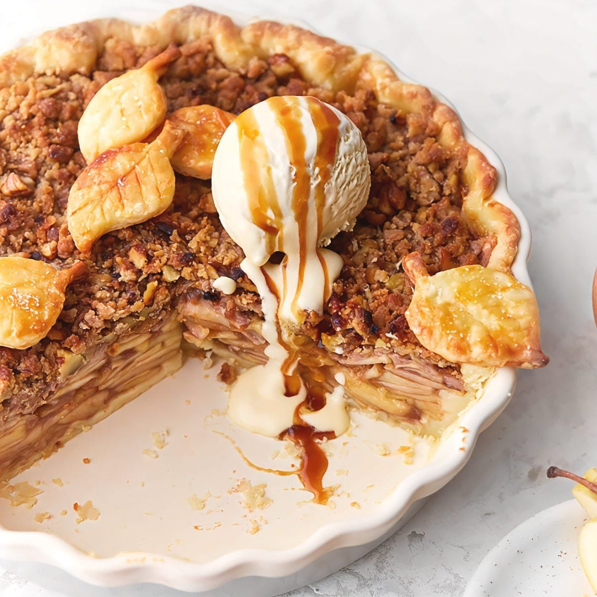 A freshly baked apple walnut crumble pie with leaf-shaped pastry cutouts, topped with a scoop of melting vanilla ice cream and caramel drizzle, making a delicious addition to Thanksgiving potluck desserts.