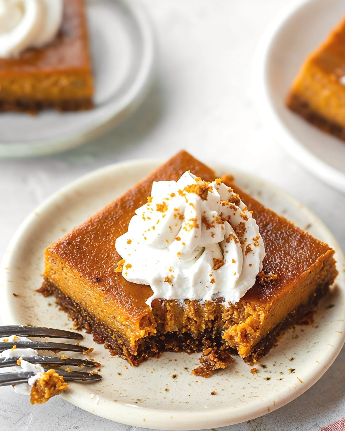 A square slice of easy pumpkin pie bar on a graham cracker crust, topped with whipped cream and fine cookie crumbs, served on a small plate for Thanksgiving potluck desserts.