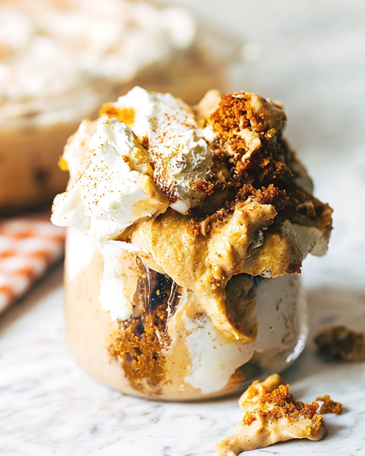 A glass cup layered with creamy pumpkin bread pudding, whipped cream, spiced crumb topping, and a dust of cinnamon, ideal for Thanksgiving potluck desserts.