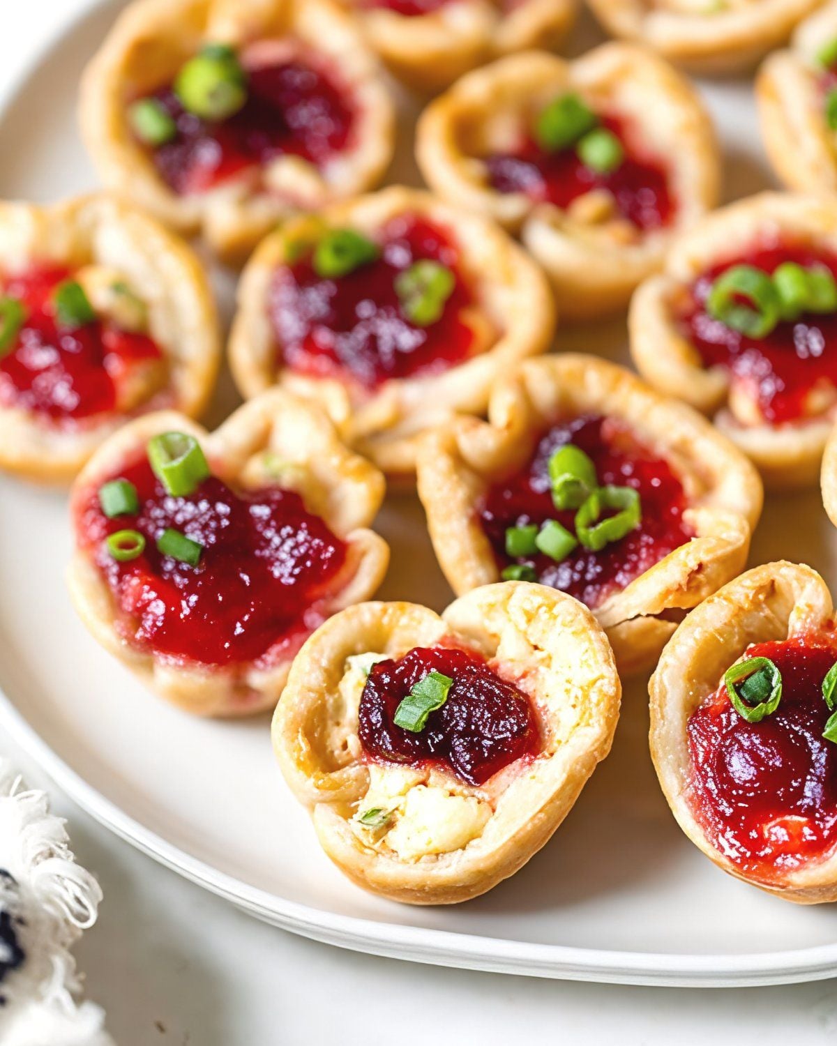 Golden, flaky pastry cups filled with cream cheese, vibrant red cranberry sauce, and fresh green scallions are arranged on a plate as delicious Thanksgiving Snacks Appetizers.