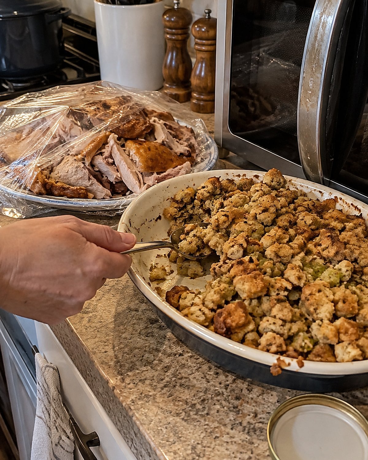 A shallow dish of stuffing sits beside a covered plate of turkey near the microwave.