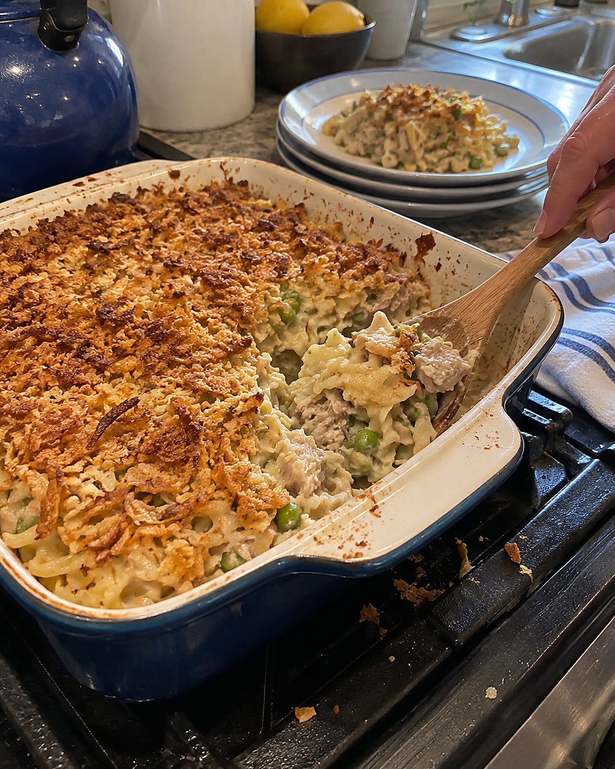 A worn casserole dish of tuna noodles sits on a stovetop, with one corner of its topping disturbed.