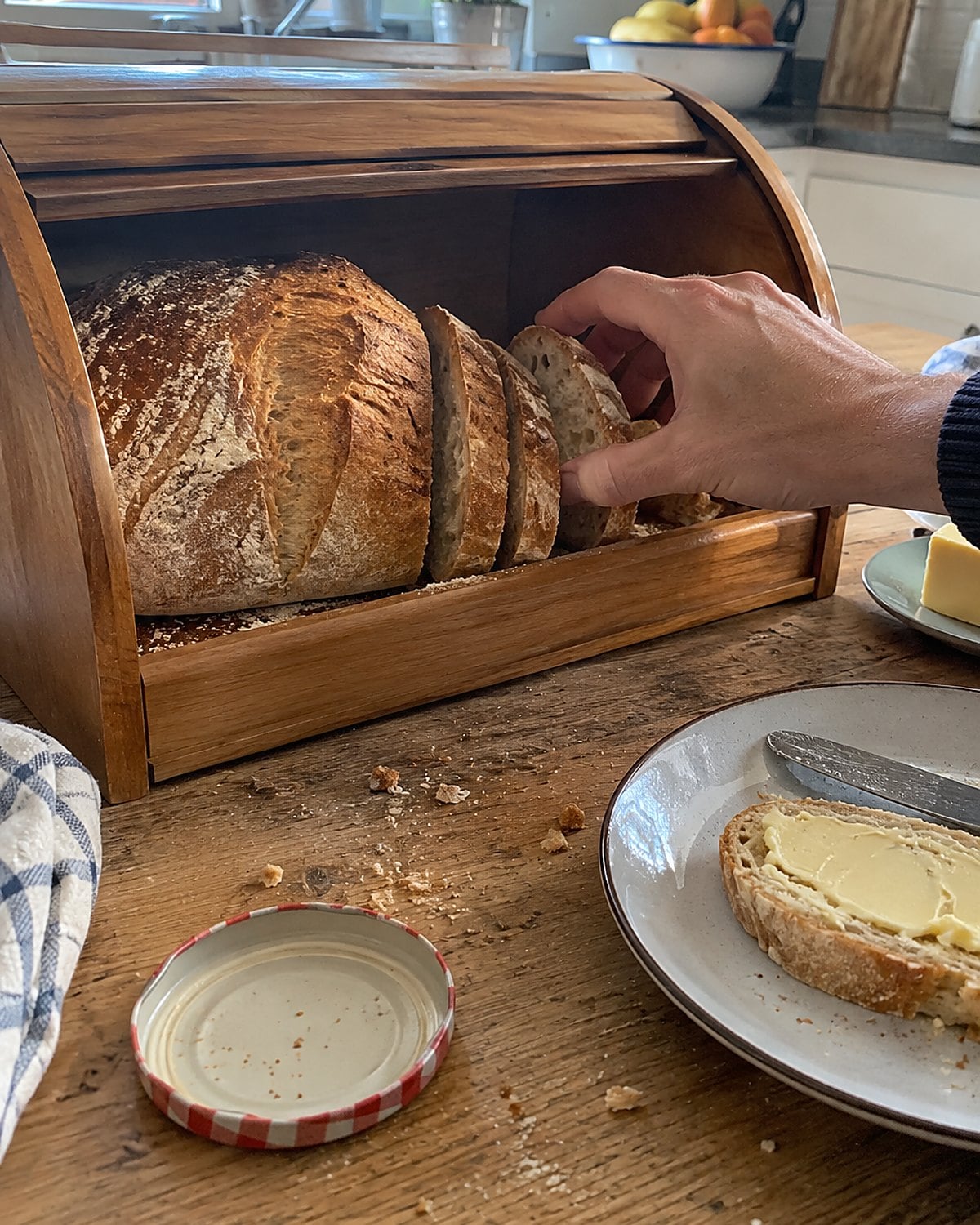 An open bread box on a kitchen table holds a partly sliced loaf and a few scattered crumbs.