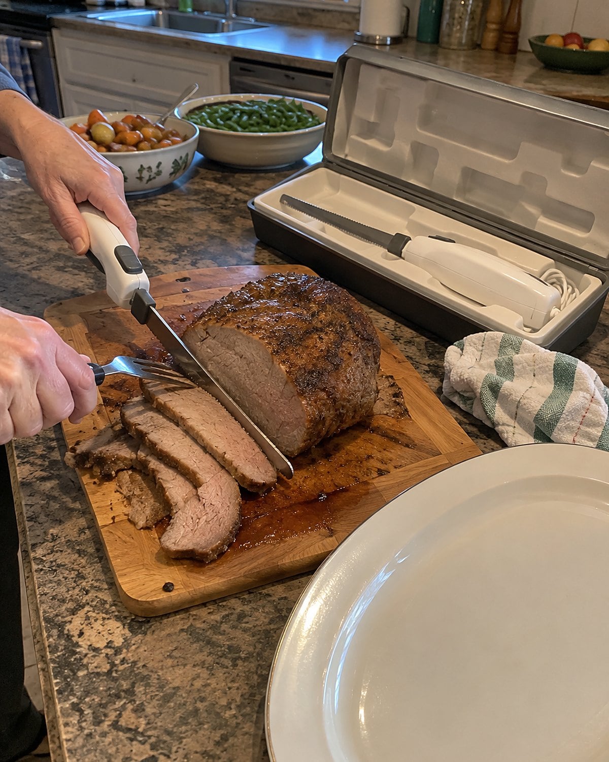 An electric knife rests in its open fitted box beside a sliced roast and a plain serving platter.