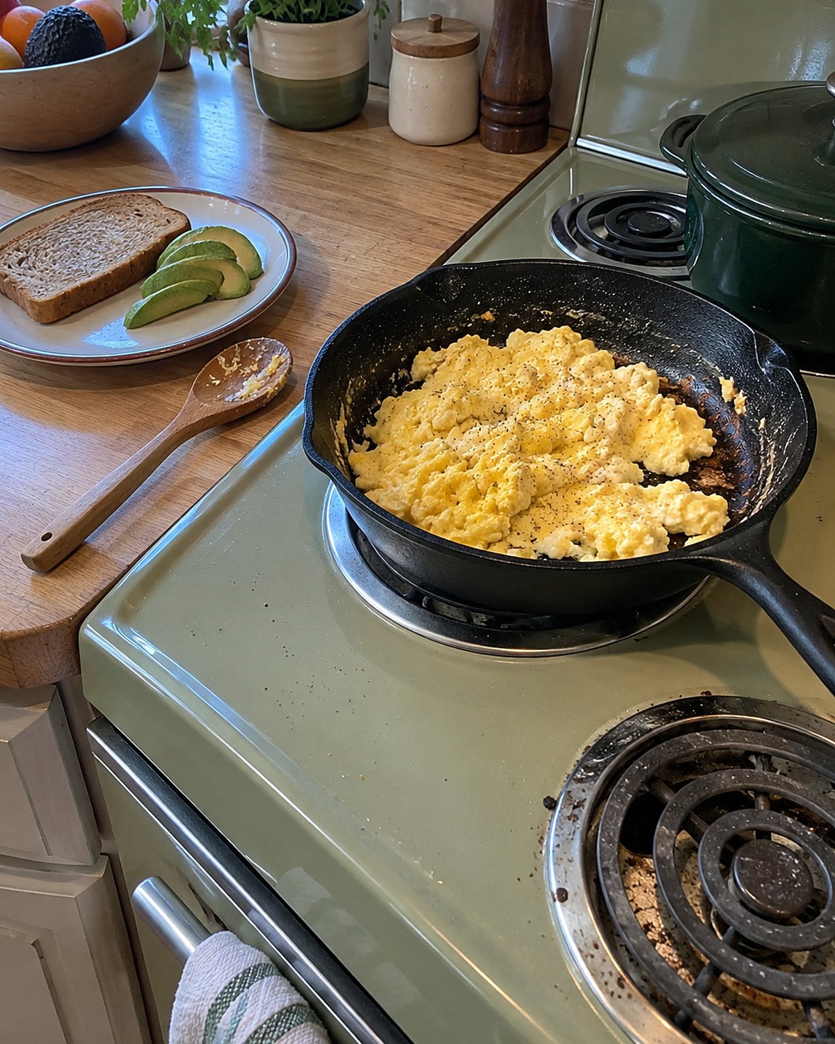 An avocado-green electric range holds a skillet of scrambled eggs, with one stained drip bowl visible beneath a coil.