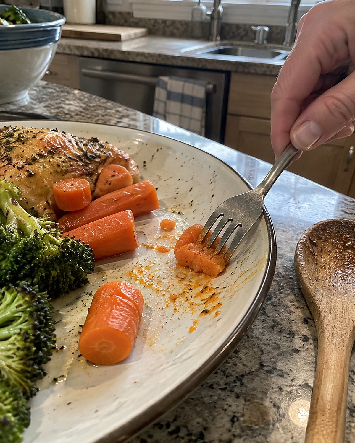A fork pressing a cooked carrot piece against a plate on the kitchen counter.