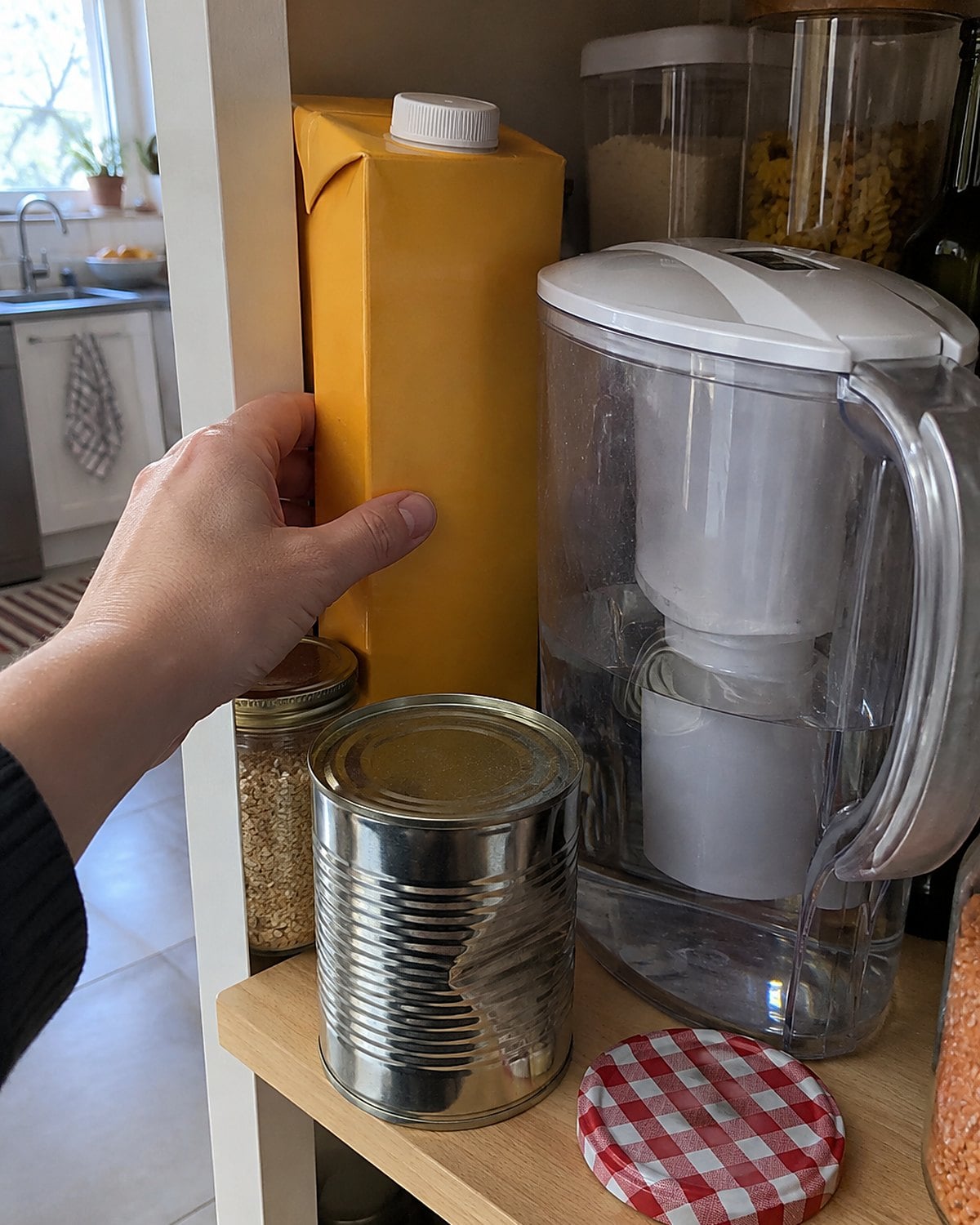 A juice box tucked behind a water pitcher on a crowded pantry shelf.