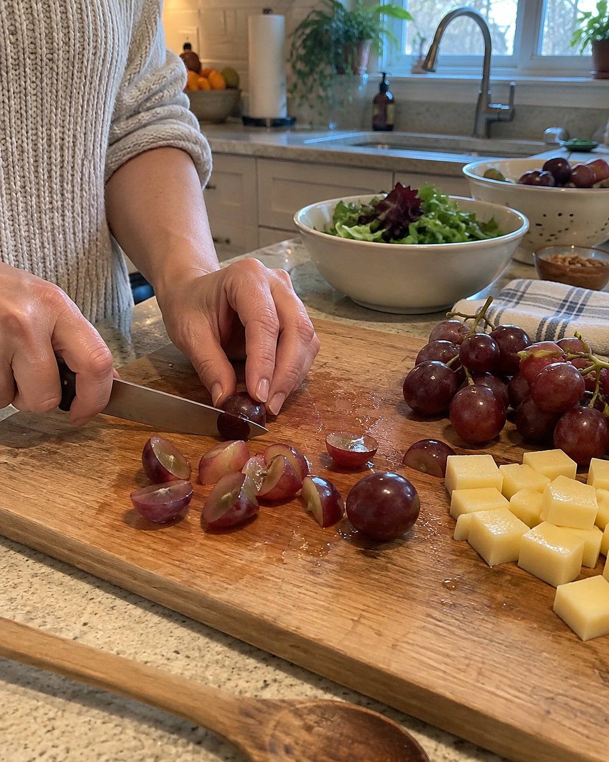 Hands quartering grapes lengthwise on a worn cutting board beside cheese cubes.