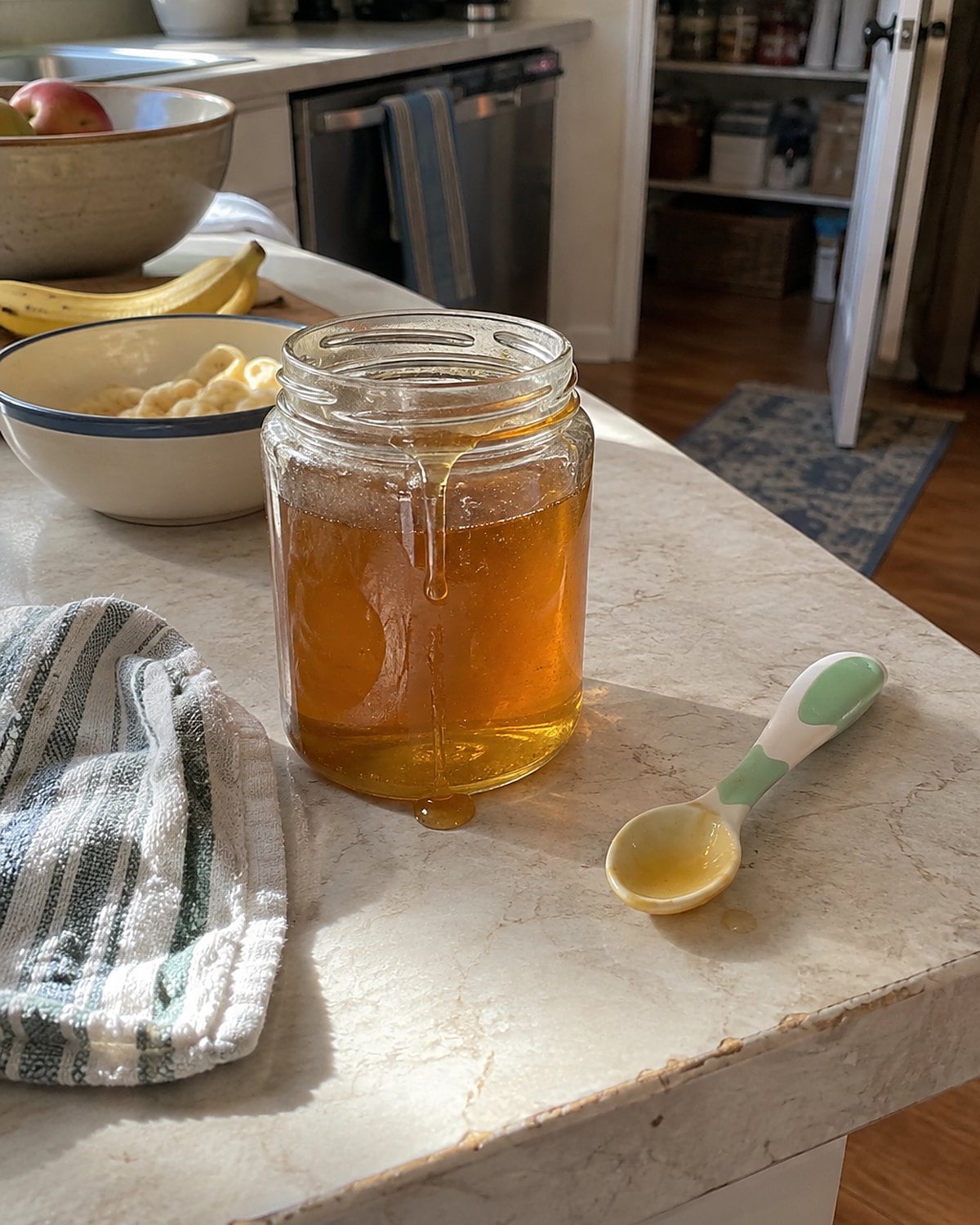 A small honey jar beside a baby spoon and an open pantry door in a home kitchen.