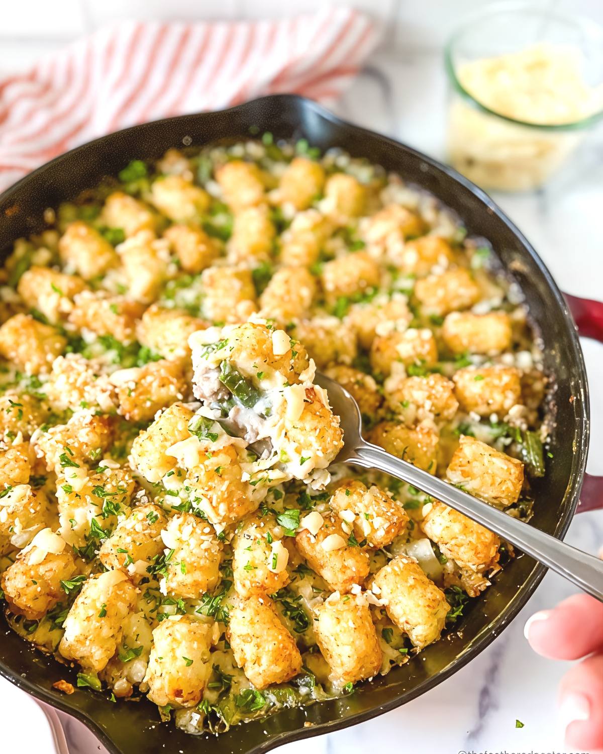 A serving spoon scoops into a cast-iron skillet filled with a turkey, green bean, and tater tot casserole garnished with fresh herbs and chopped almonds.