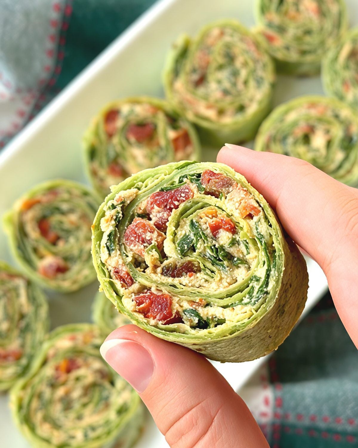 A hand holding a green spinach wrap pinwheel filled with tofu and sun-dried tomatoes, served as one of the best vegan Thanksgiving appetizers.