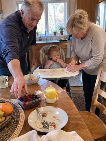 Early morning at a grandchildโs high chair at the kitchen table, a couple in their sixties wiping spilled cereal and reaching for a carton of berries, nobody posing.