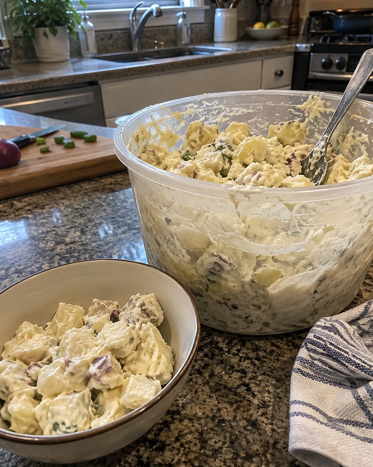 A large tub of potato salad beside a small serving bowl on a kitchen counter.