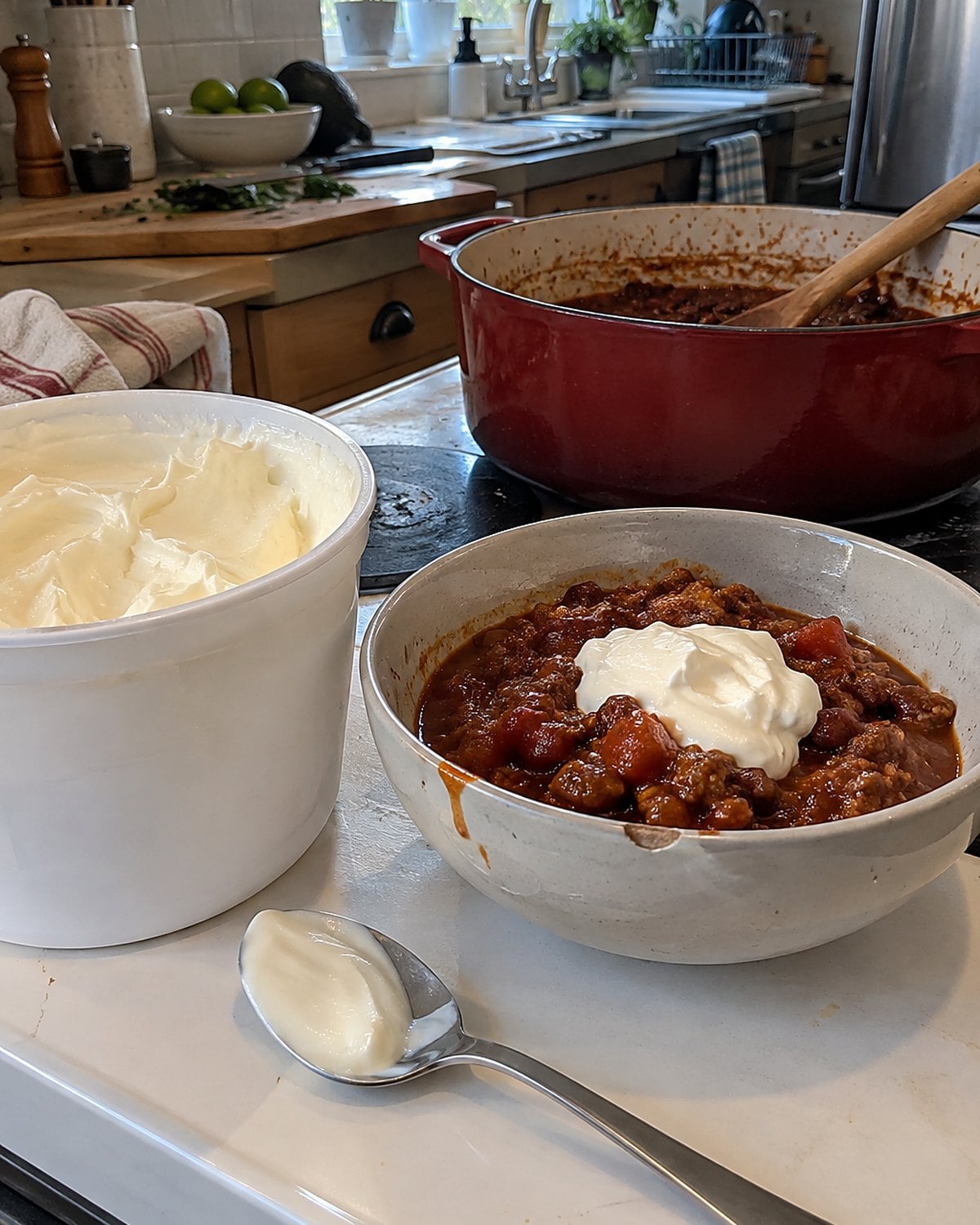 A wide sour cream tub and a clean spoon beside a bowl of chili on the stove.