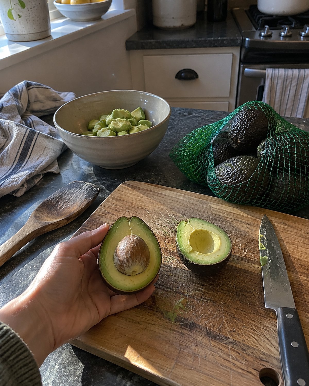 A mesh sack of avocados beside one cut avocado on a worn cutting board.