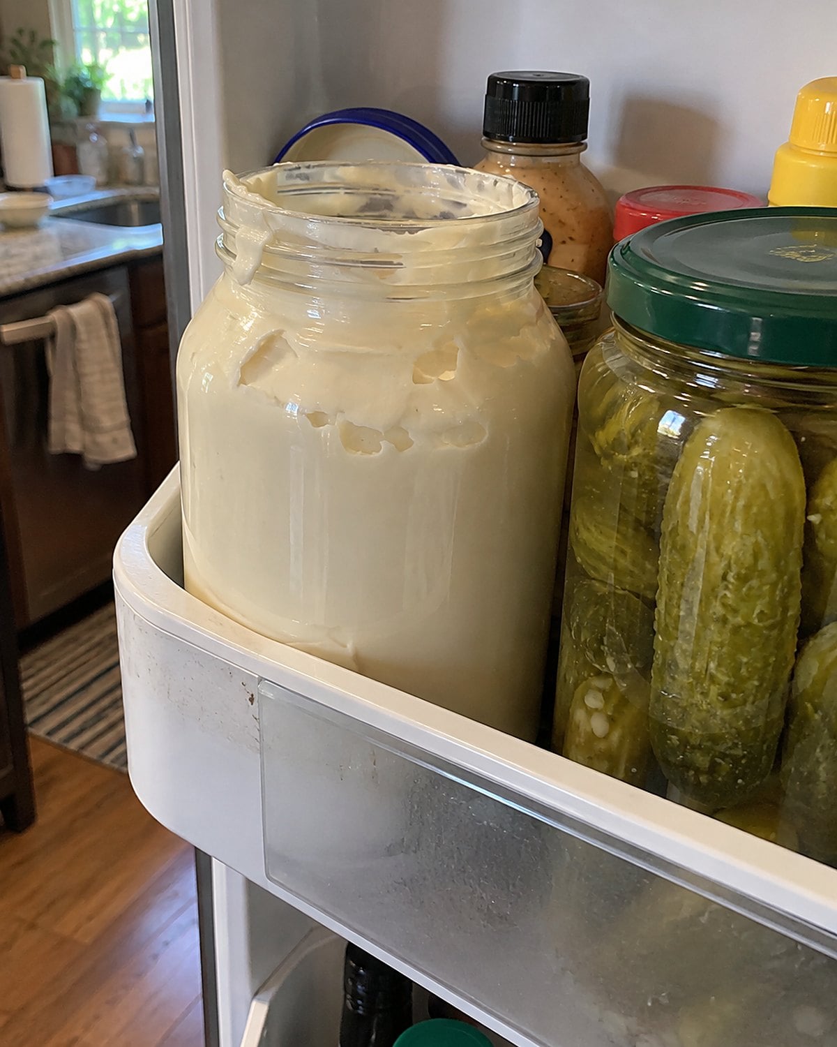 An opened jumbo mayonnaise jar beside pickles on a crowded refrigerator door shelf.