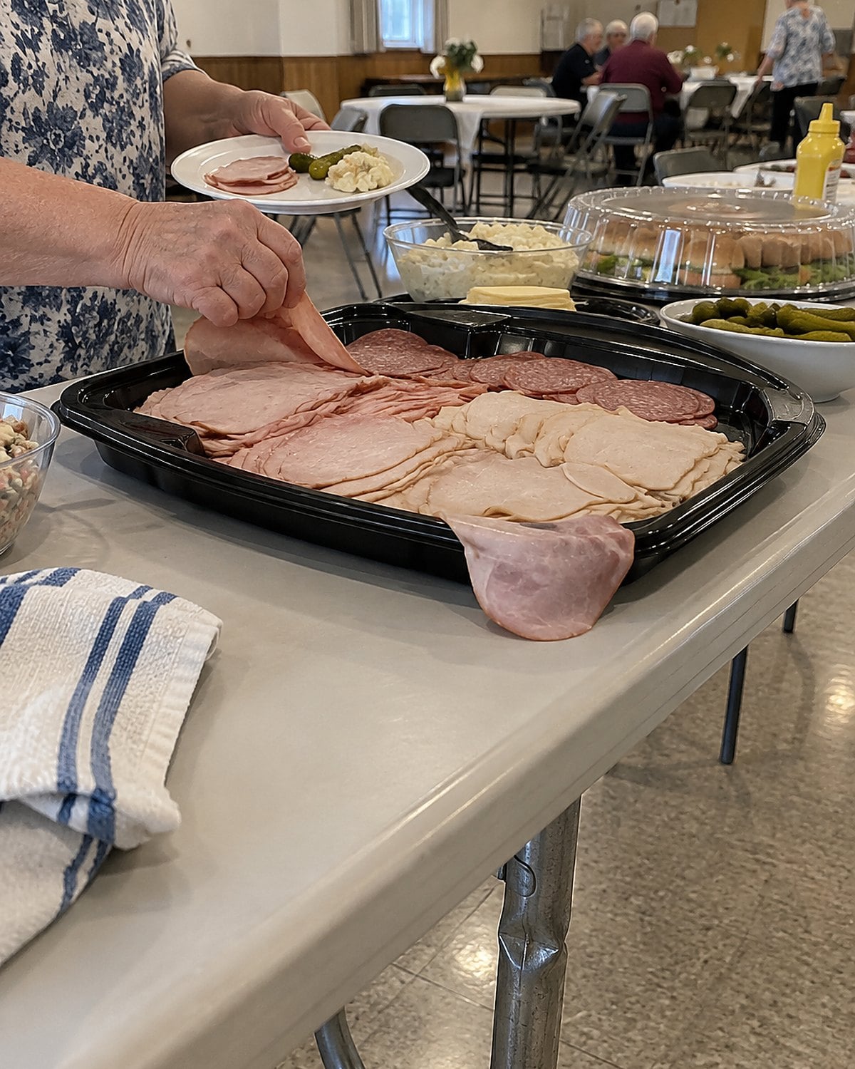 A partly used deli meat tray beside a covered serving plate on a church hall table.